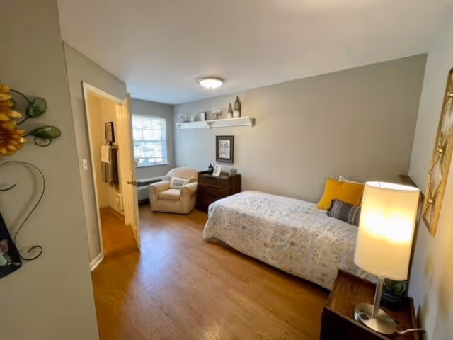 A cozy bedroom with a single bed covered in a floral bedspread, a yellow pillow, and a gray pillow. There is a wooden nightstand with a lamp on it next to the bed. Across from the bed is a wooden dresser with decorative items and a framed picture above it. A comfortable armchair is placed near a window with white blinds. The walls are painted light gray, and the floor is wooden. A door to a bathroom is open on the left side of the room.