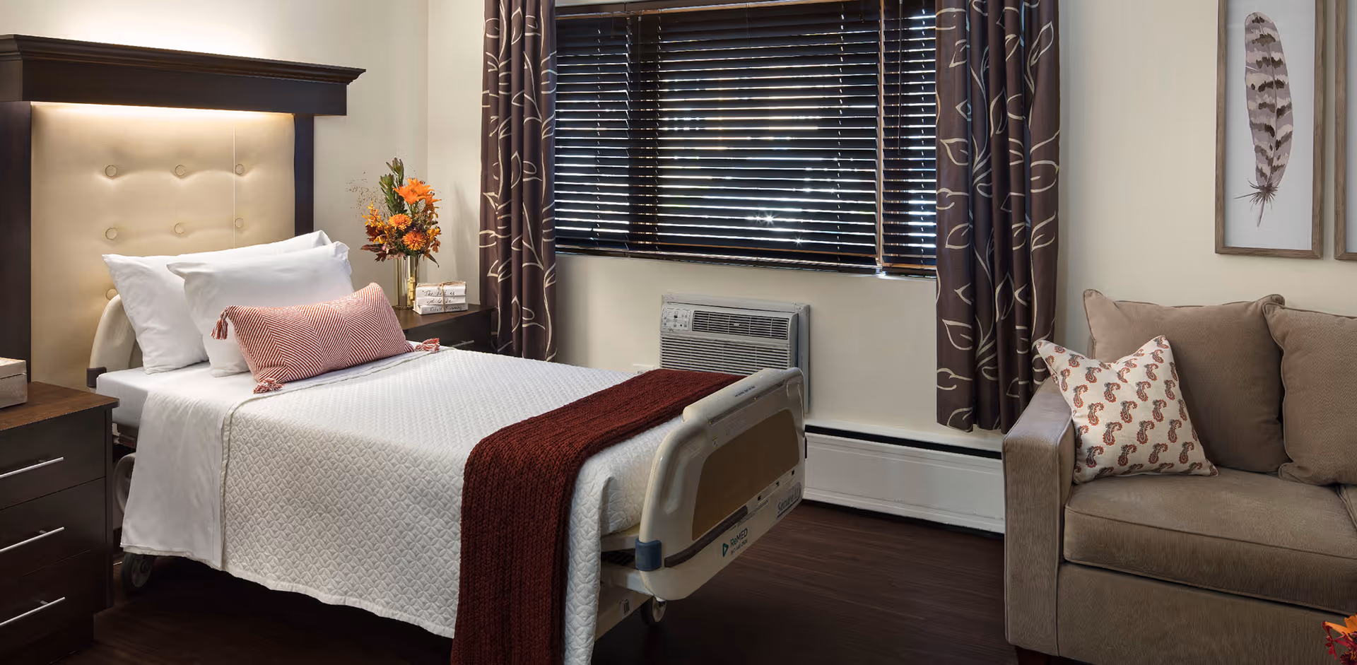 A neatly made hospital-style bed with white bedding and a red throw blanket in a cozy room. The bed has a padded headboard and is positioned next to a window with dark blinds and patterned curtains. There is a nightstand with a vase of flowers and decorative blocks, and a beige sofa with patterned pillows is visible on the right side of the image. The room has wooden flooring and framed feather artwork on the wall.