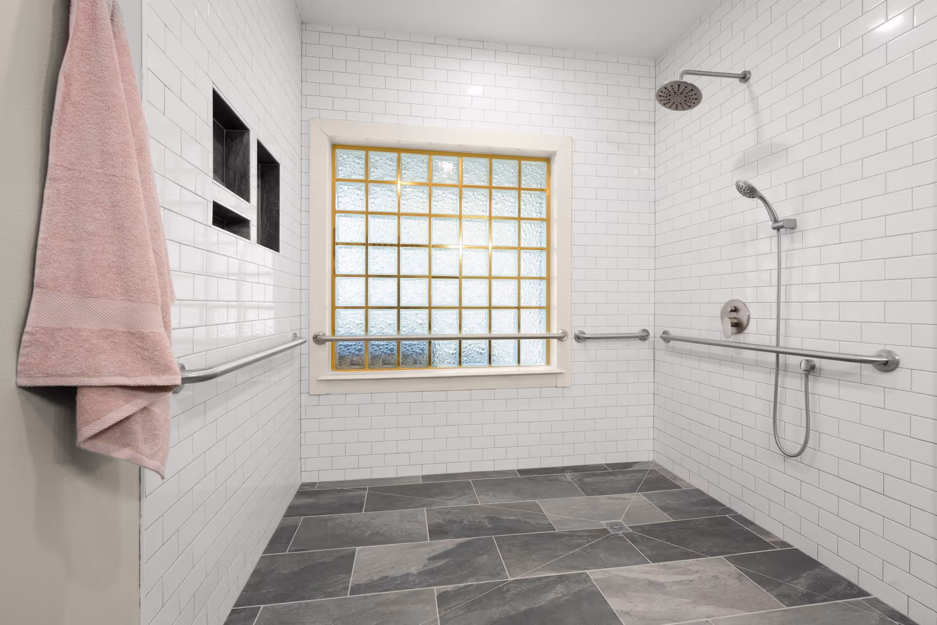 A spacious, accessible shower area with white subway tile walls and dark gray floor tiles. The shower features a rainfall showerhead, a handheld showerhead, multiple grab bars, and built-in wall niches for storage. A pink towel hangs on a hook on the left wall, and a frosted glass block window allows natural light into the space.