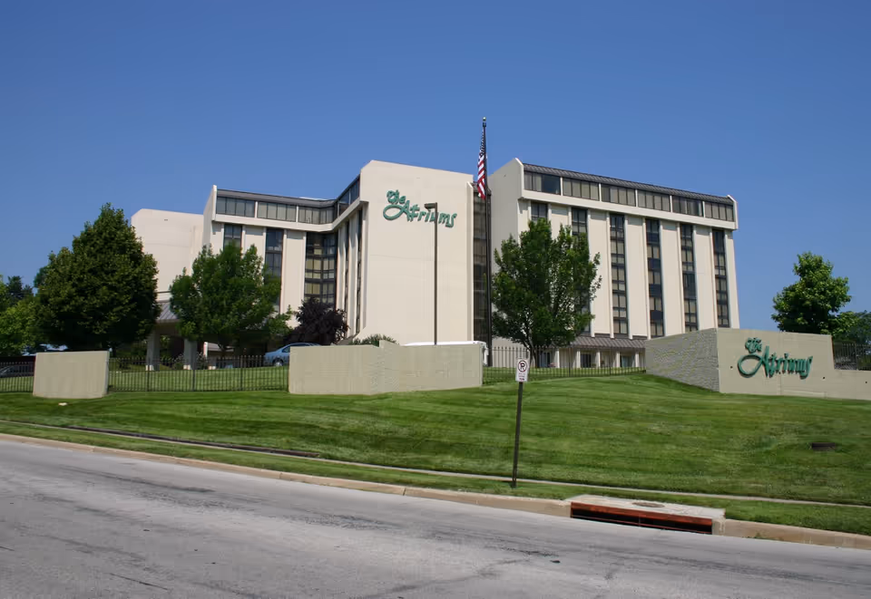 Exterior view of The Atriums Senior Living Community building with multiple floors, surrounded by green grass and trees under a clear blue sky. An American flag is visible in front of the building.