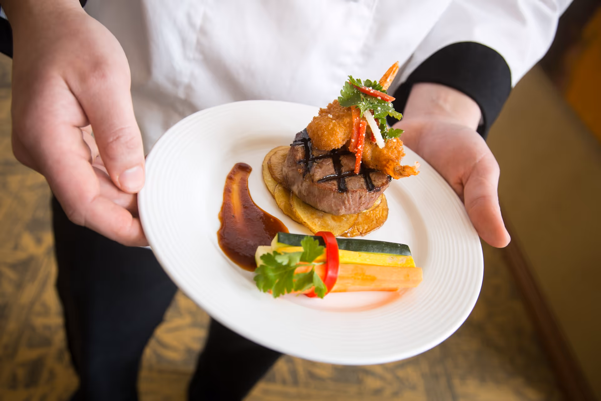 A person wearing a white chef's jacket and black pants holding a white plate with a gourmet dish consisting of grilled steak topped with fried shrimp and garnished with herbs, served with a side of colorful vegetable sticks and a smear of sauce.