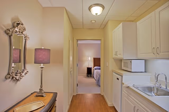 View down a narrow hallway with a small kitchen area on the right featuring white cabinets, a microwave, and a sink. On the left, there is a decorative mirror above a wooden console table with a lamp. At the end of the hallway, a bedroom is visible with a bed, nightstand, and lamp.