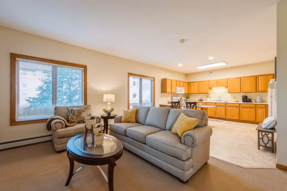 A bright and spacious living room and kitchen area in a senior living facility. The living room features a gray sofa with yellow pillows, a matching armchair with a patterned cushion, a round wooden coffee table with decorative items, and a side table with a lamp and plant. The kitchen has wooden cabinets, a white microwave, stove, refrigerator, and a small dining table with chairs. Large windows with blinds allow natural light to fill the space.