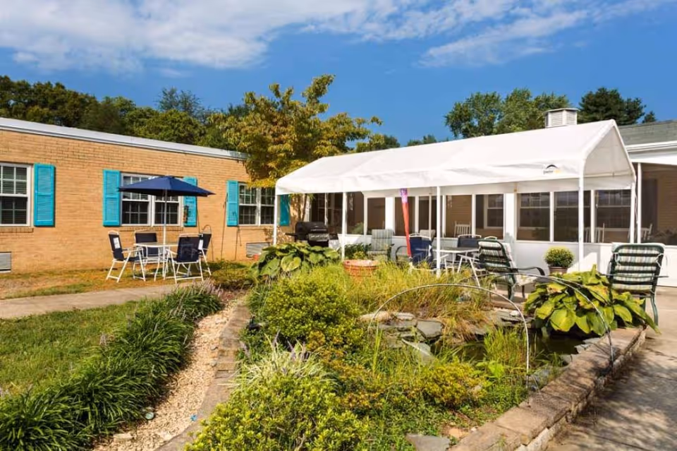 Outdoor courtyard area of a senior living facility with a garden pond surrounded by plants and greenery. There are several chairs and tables with umbrellas, and a white canopy providing shade. The building has beige brick walls and blue window shutters under a partly cloudy sky.