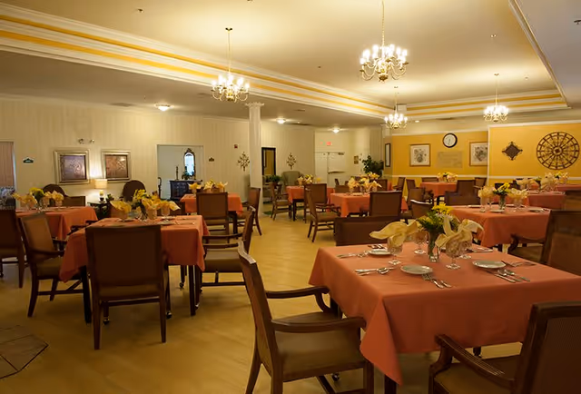 A spacious dining room with multiple tables covered in orange tablecloths, each set with plates, silverware, glasses, and yellow napkins. The room features wooden chairs, chandeliers hanging from the ceiling, framed artwork on the walls, and a warm yellow and beige color scheme.