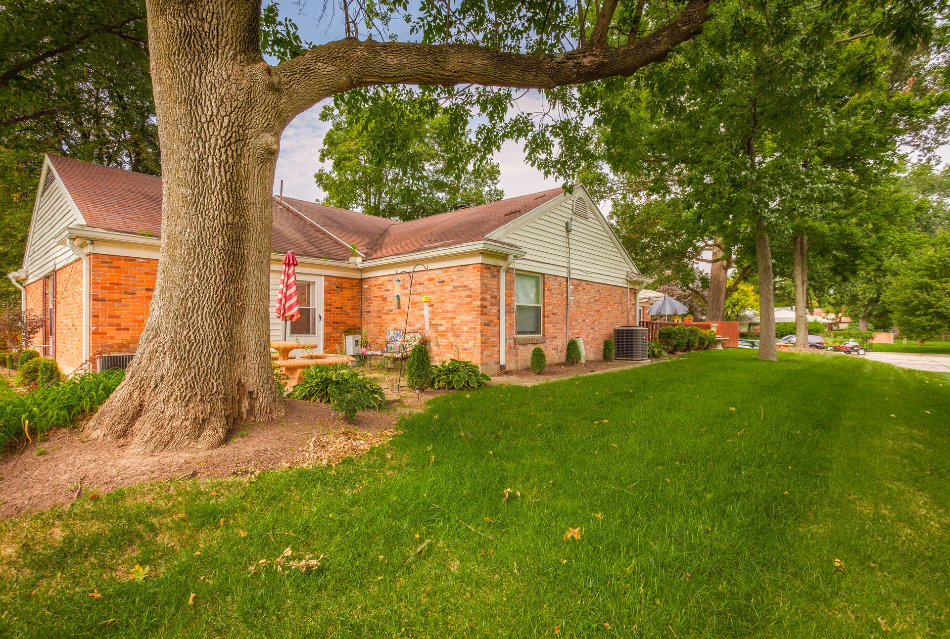 Exterior view of a single-story brick building with a large tree in the foreground and green grass surrounding the area. There is a small patio with a table, chairs, and a red and white striped umbrella near the building.