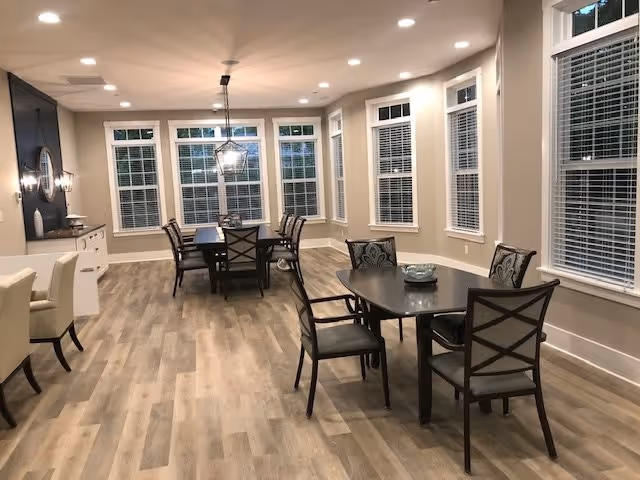 A spacious dining area with multiple tables and chairs arranged neatly. The room features large windows with white blinds, light-colored walls, and wood-patterned flooring. Ceiling lights and a hanging light fixture illuminate the space.