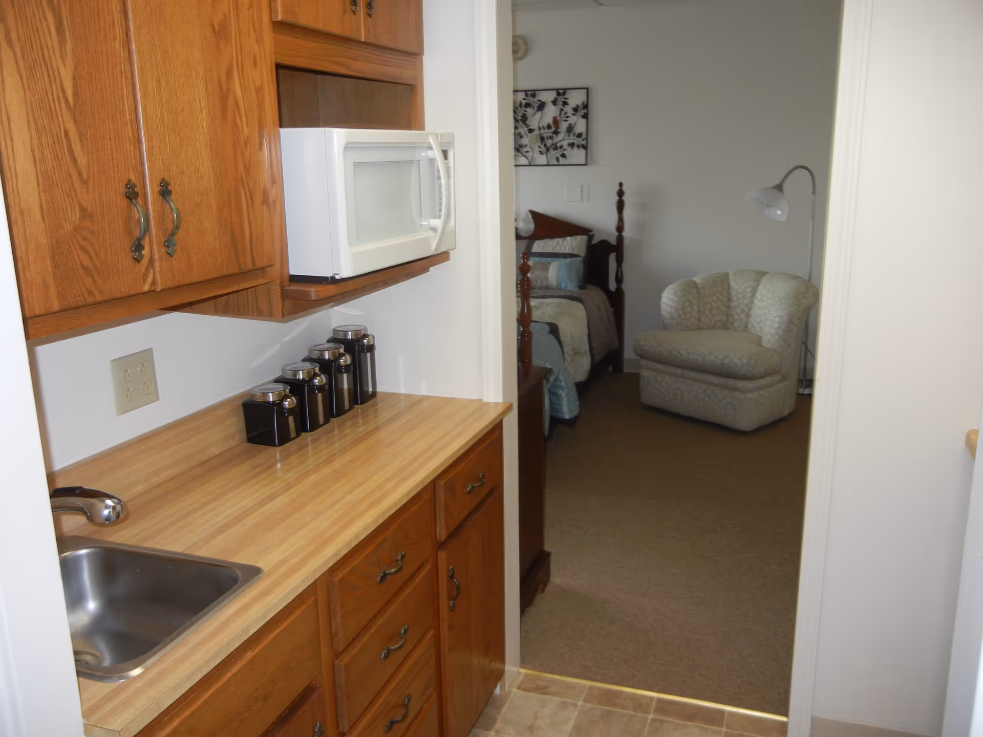 View of a small kitchen area with wooden cabinets, a countertop with a sink, a microwave, and a set of black canisters. Through an open doorway, a bedroom is visible with a bed, a cushioned armchair, a floor lamp, and a piece of wall art.
