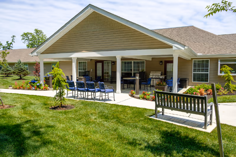 Outdoor covered patio area at a senior living facility with several blue cushioned chairs arranged around tables, a grill, and a bench on a concrete pathway surrounded by green grass and landscaped plants.
