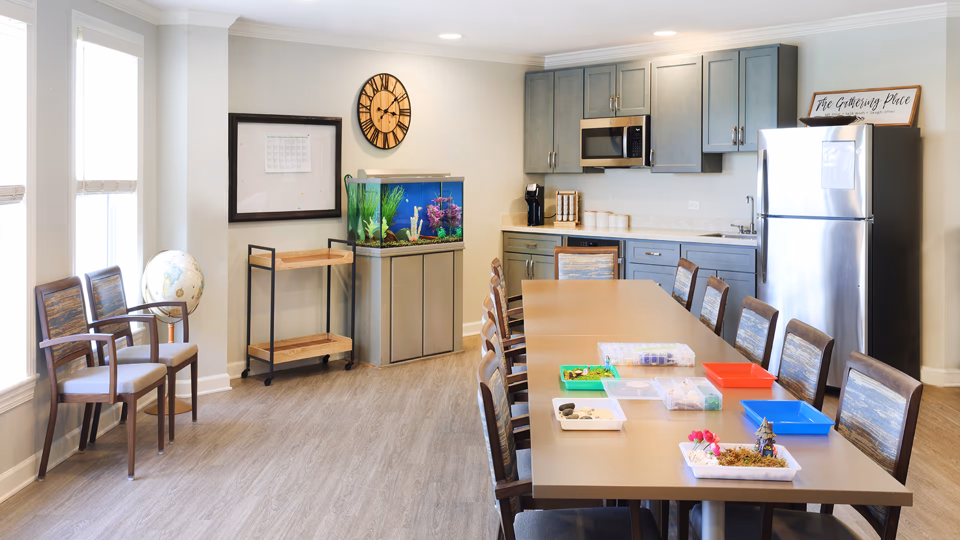 A bright room with a long table surrounded by chairs, some craft supplies on the table, a kitchen area with gray cabinets, a microwave, and a refrigerator. There is a fish tank on a stand against the wall, a globe on a stand near two chairs by the windows, and a wall clock above the fish tank. A sign on top of the refrigerator reads 'The Gathering Place.'