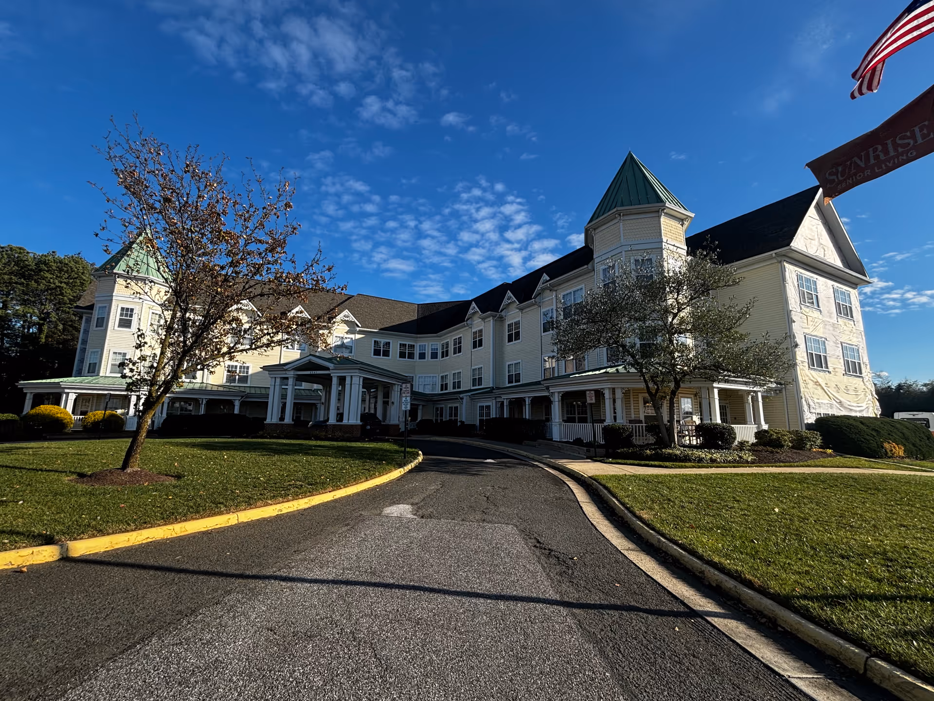 Exterior view of a large, multi-story senior living facility building with light yellow siding, green roofs on turret-like structures, and a covered entrance driveway. There are trees and well-maintained lawns on either side of the driveway, and a flagpole with an American flag and a Sunrise Senior Living banner is visible on the right side under a blue sky with scattered clouds.
