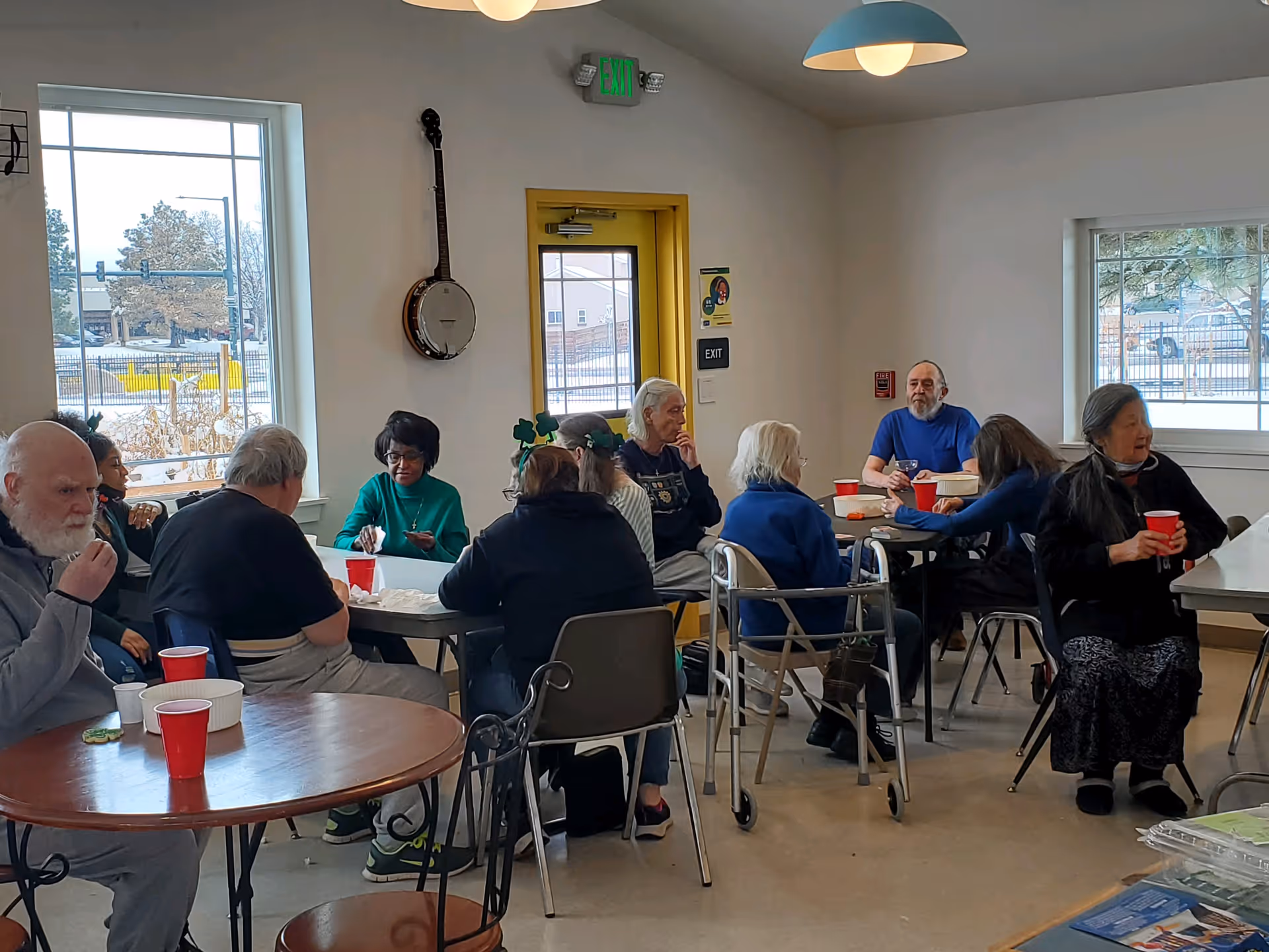 A group of elderly people sitting around tables in a bright room with large windows, drinking from red cups and engaging in conversation. The room has a yellow door with an exit sign above it, a banjo hanging on the wall, and pendant lights hanging from the ceiling.