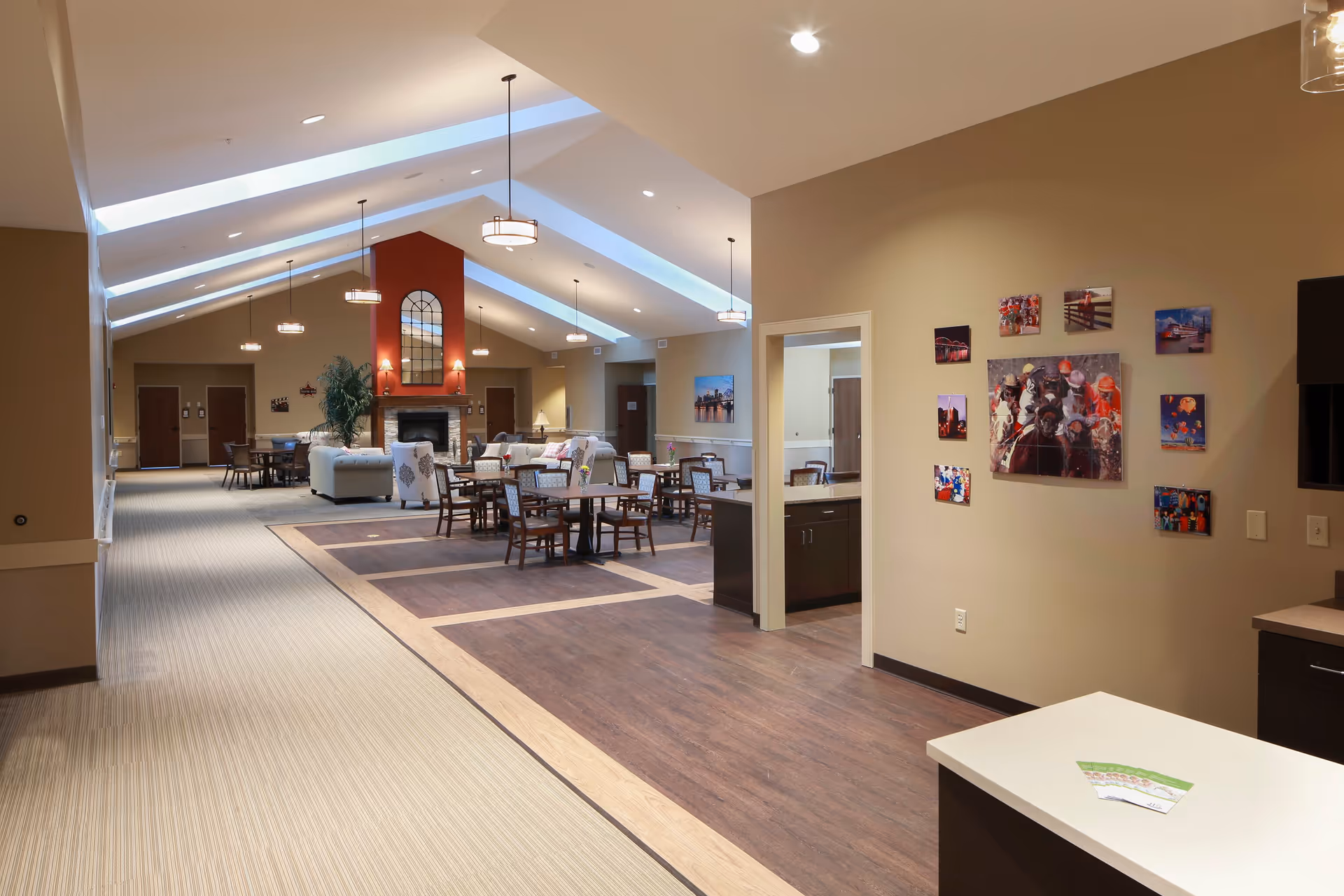 A spacious, well-lit common area in a senior living facility with high vaulted ceilings and hanging pendant lights. The room features multiple seating arrangements including tables with chairs and sofas around a central fireplace with a large mirror above it. The walls are painted beige, and there are several colorful pictures displayed on one wall near a small kitchenette area.