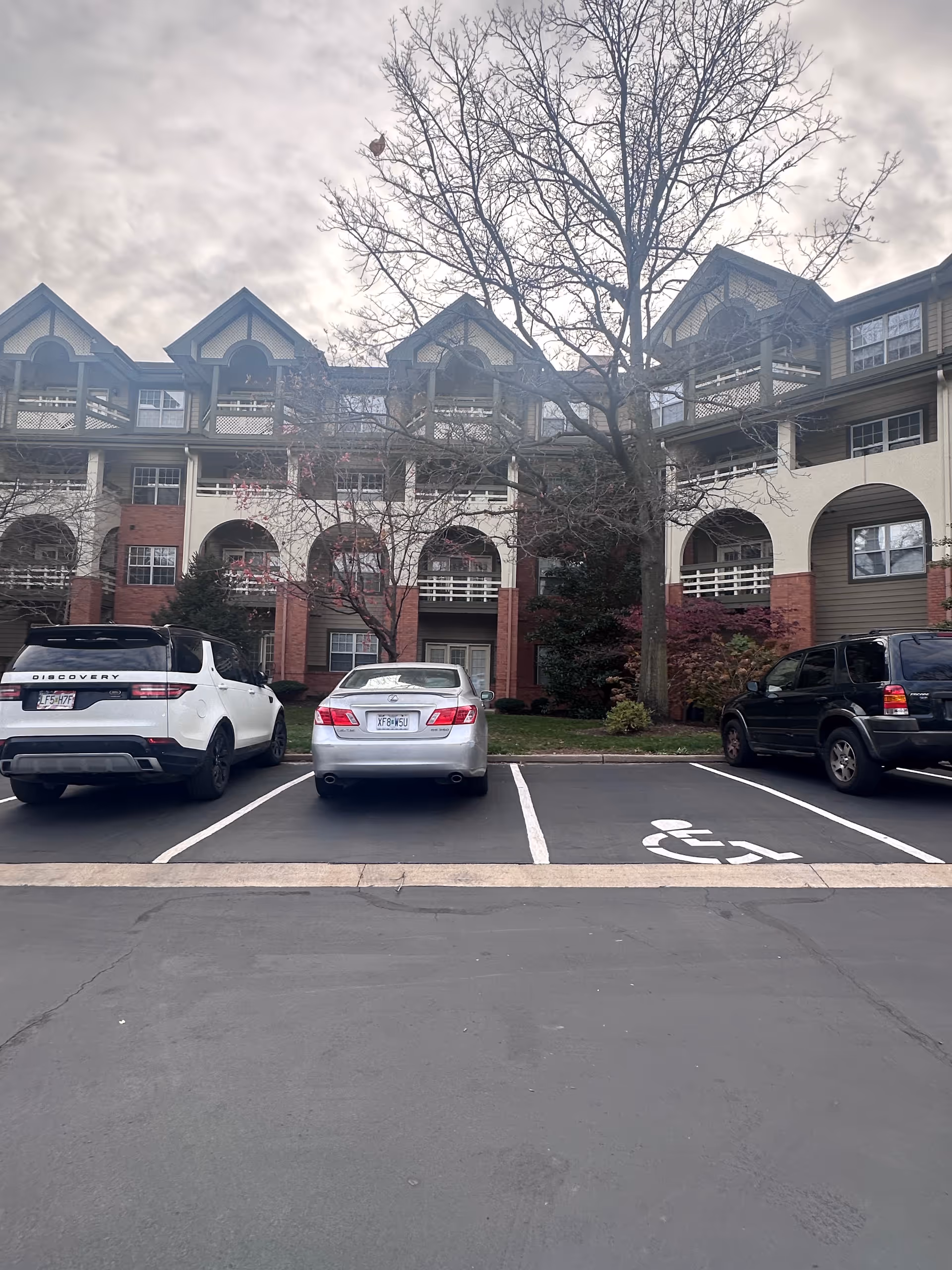 Parking lot in front of a multi-story residential building with arched balconies and a tree without leaves. Three cars are parked in the lot, including one in a handicapped parking space.