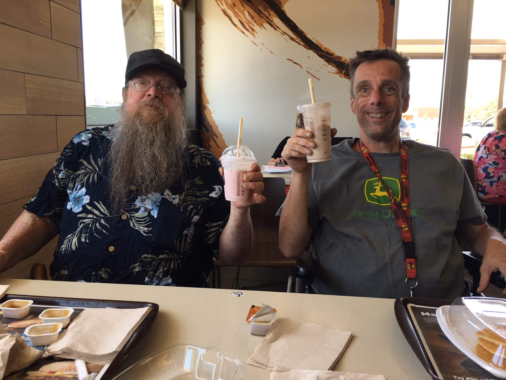 Two men sitting at a table inside a dining area, each holding a milkshake and smiling. One man has a long beard, glasses, and a black floral shirt, while the other wears a John Deere t-shirt and a lanyard. There are trays with napkins and sauce containers on the table, and a window with sunlight in the background.