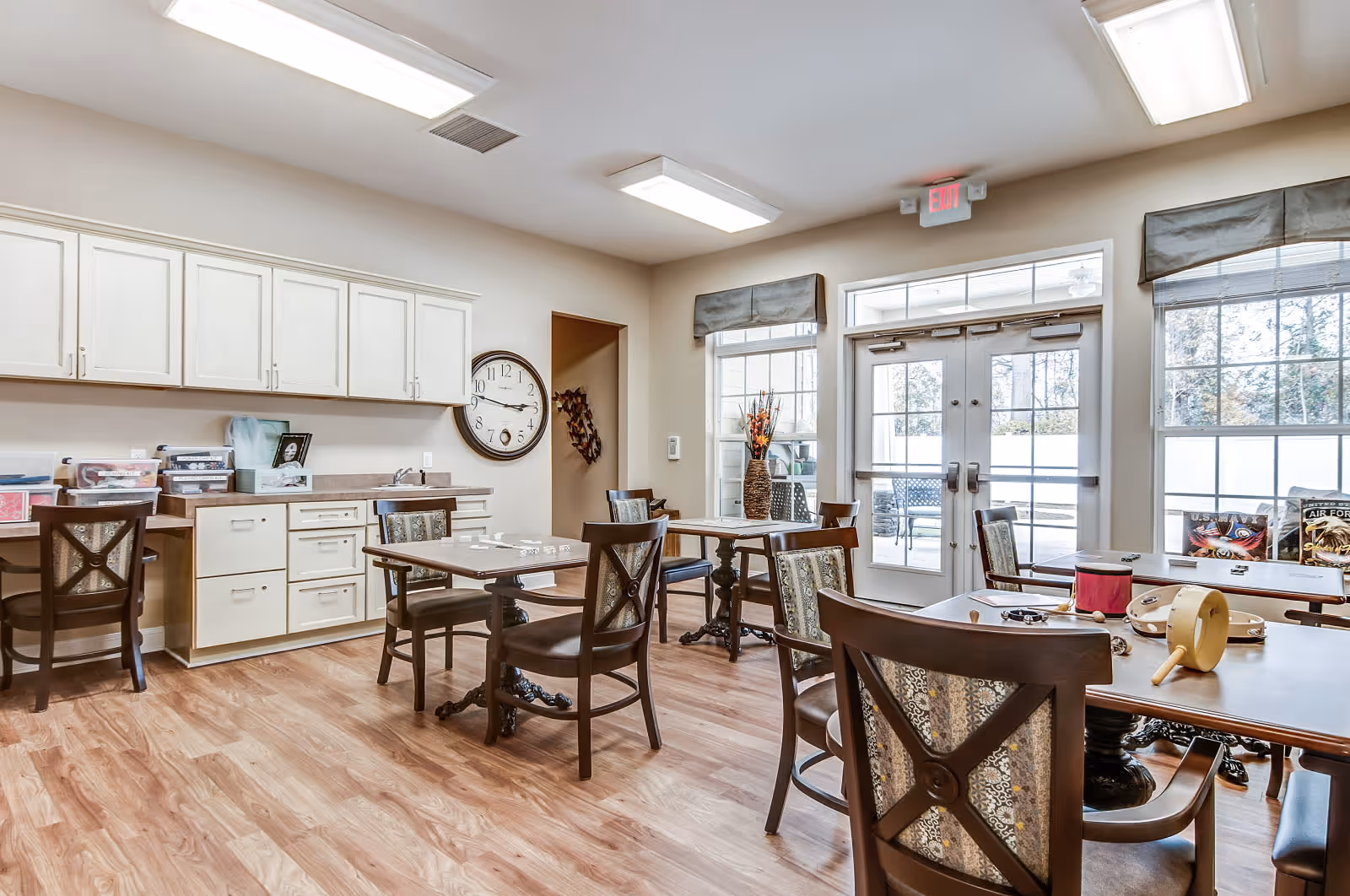 A bright and clean activity room with wooden flooring, several tables and chairs arranged for group activities, a large wall clock, white cabinets, and double glass doors leading outside. The room has large windows with valances and various small items on the tables.