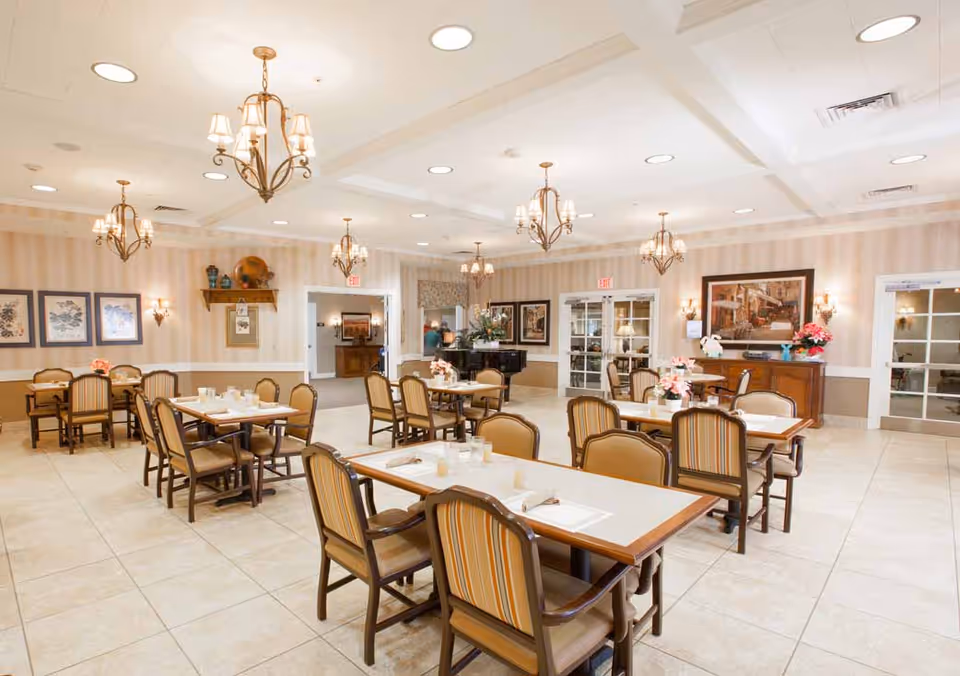 A spacious dining room with multiple tables and chairs arranged neatly. The room features beige tiled floors, soft striped wallpaper, and several chandeliers hanging from the ceiling. There are framed pictures on the walls, floral centerpieces on the tables, and a piano in the background near a set of double doors.