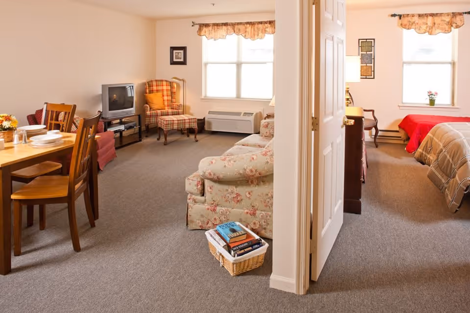 Interior view of a senior living facility apartment showing a living room and a bedroom separated by a partially open door. The living room has a floral patterned sofa, a plaid armchair with an orange pillow, a TV on a stand, and a wooden dining table with chairs set with plates and bowls. The bedroom has two beds with red and beige bedspreads, a wooden dresser, a chair, and windows with floral valances letting in natural light.