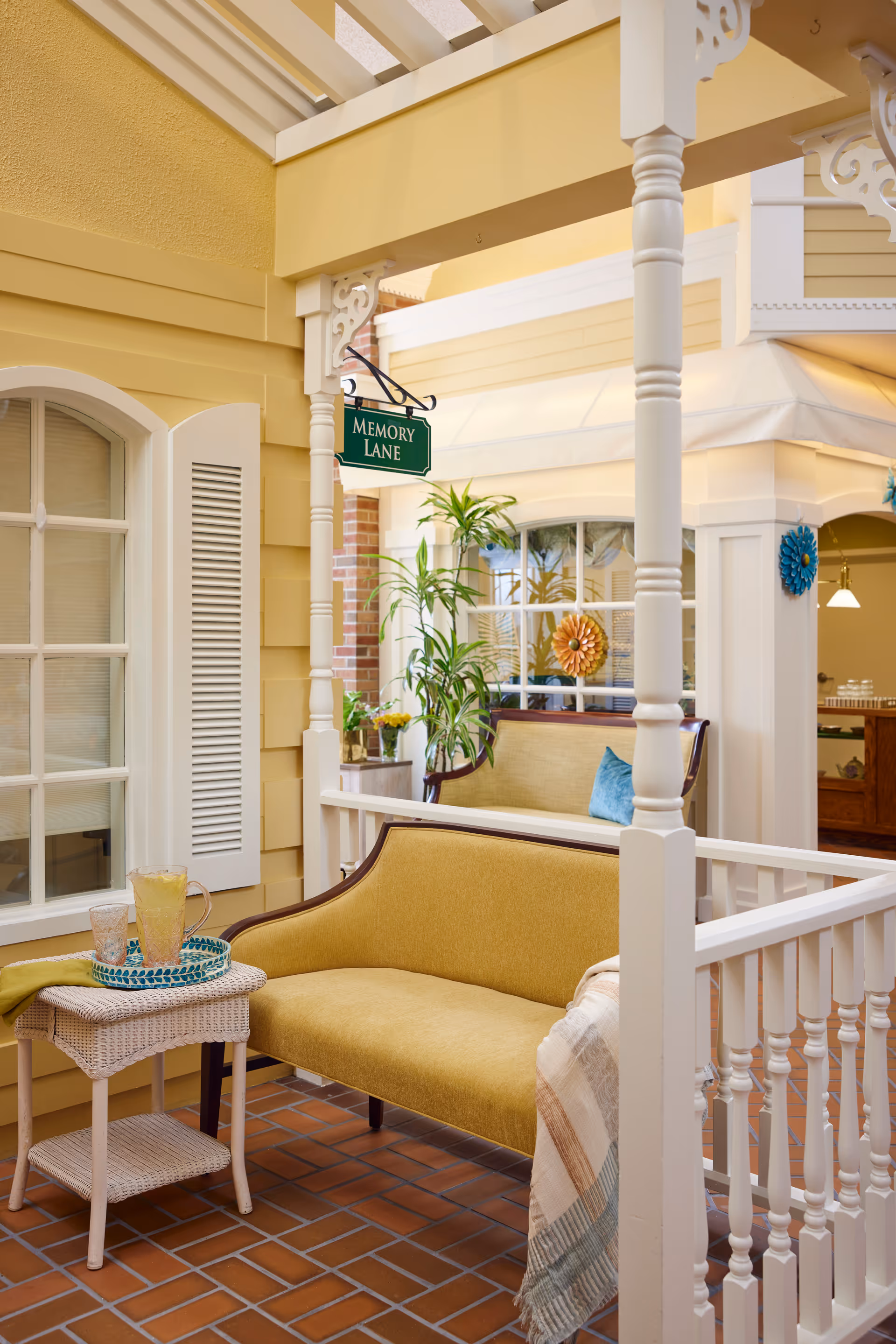 A cozy indoor seating area with yellow walls and white trim, featuring a yellow upholstered bench, a small white wicker side table with a pitcher and glasses, potted plants, and a green sign that reads 'Memory Lane'. The floor is tiled with reddish-brown bricks, and there is a white railing in the foreground.