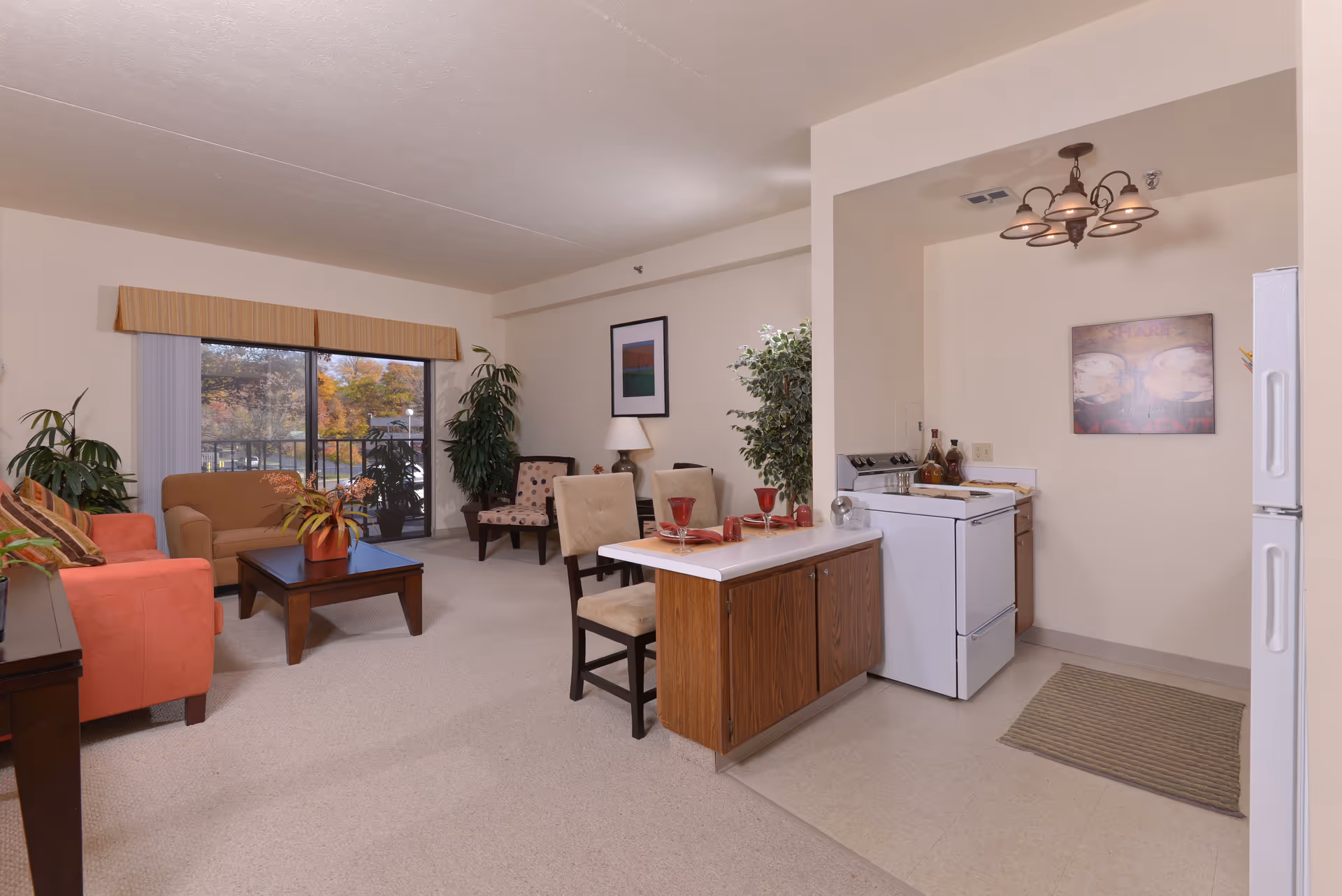 Open-plan living area with sofas and chairs around a coffee table, potted plants, sliding glass door, and a small kitchen with an island and stove.