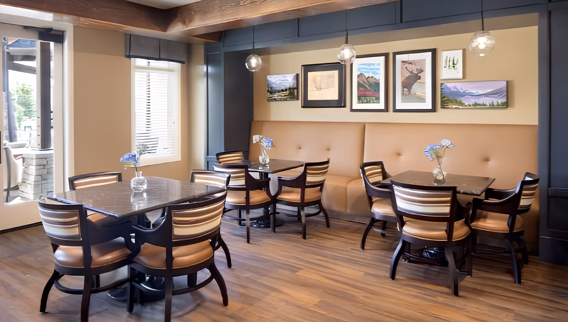 A cozy dining area with wooden floors, three tables each surrounded by four chairs with striped backs and tan seats. Each table has a small vase with blue flowers. The back wall features a cushioned bench seat and several framed artworks. Pendant lights hang from the ceiling, and there is a window and a glass door leading outside.