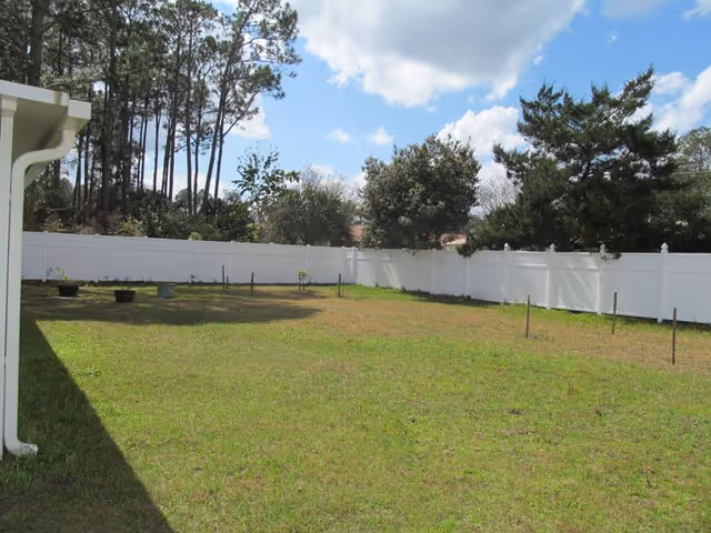 A fenced backyard area with green grass, several small plants in pots, and some trees in the background under a partly cloudy blue sky.