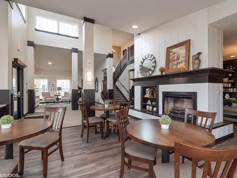 Interior view of a senior living community common area with round wooden tables and chairs, a fireplace with decorative items on the mantel, a staircase leading to an upper floor, and large windows allowing natural light to fill the space.