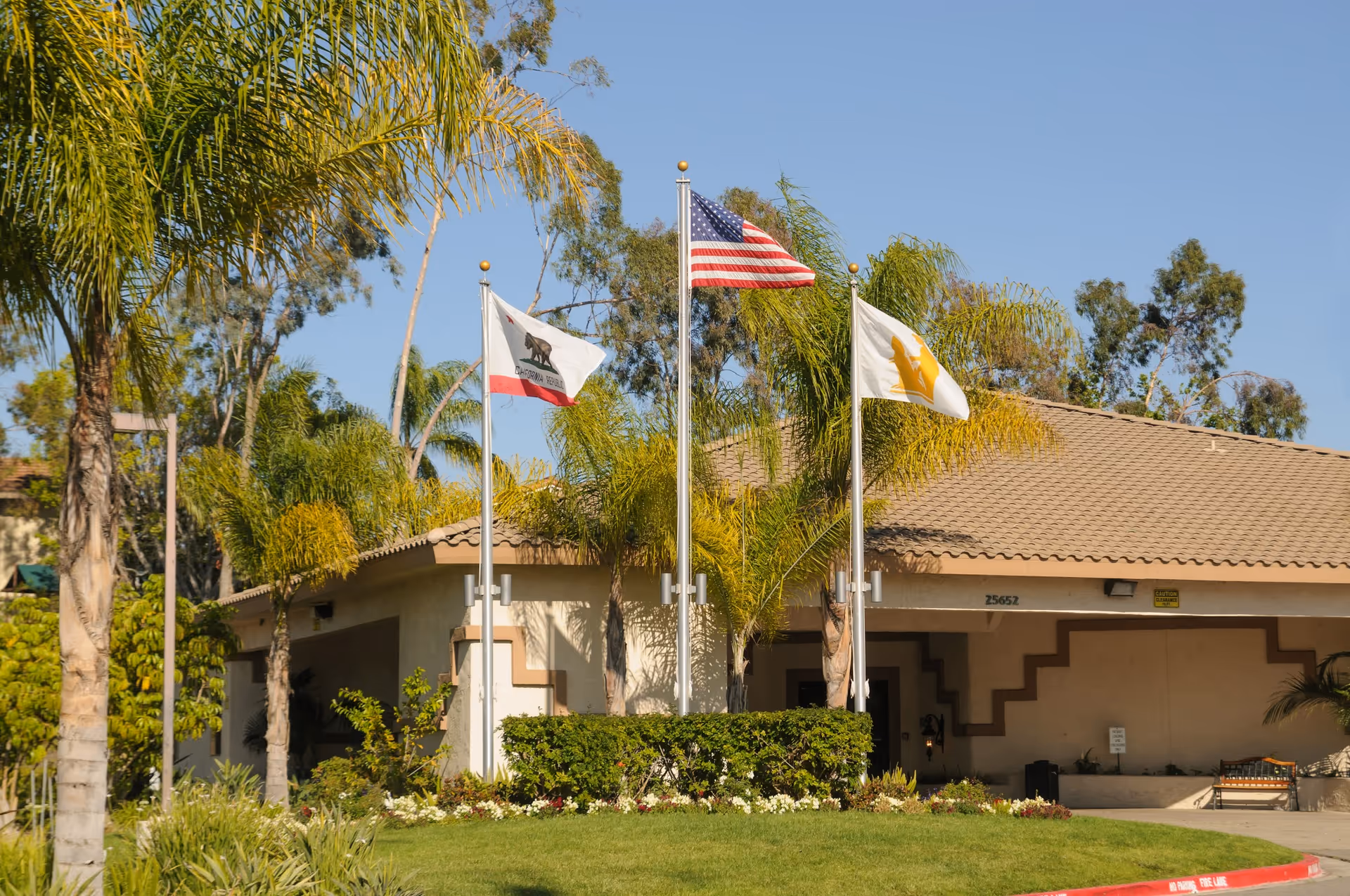 Exterior view of a single-story building with a tiled roof, surrounded by palm trees and greenery. Three flagpoles stand in front of the building, displaying the American flag in the center, the California state flag on the left, and another flag with a yellow emblem on the right. The building has the number 25652 above the entrance.