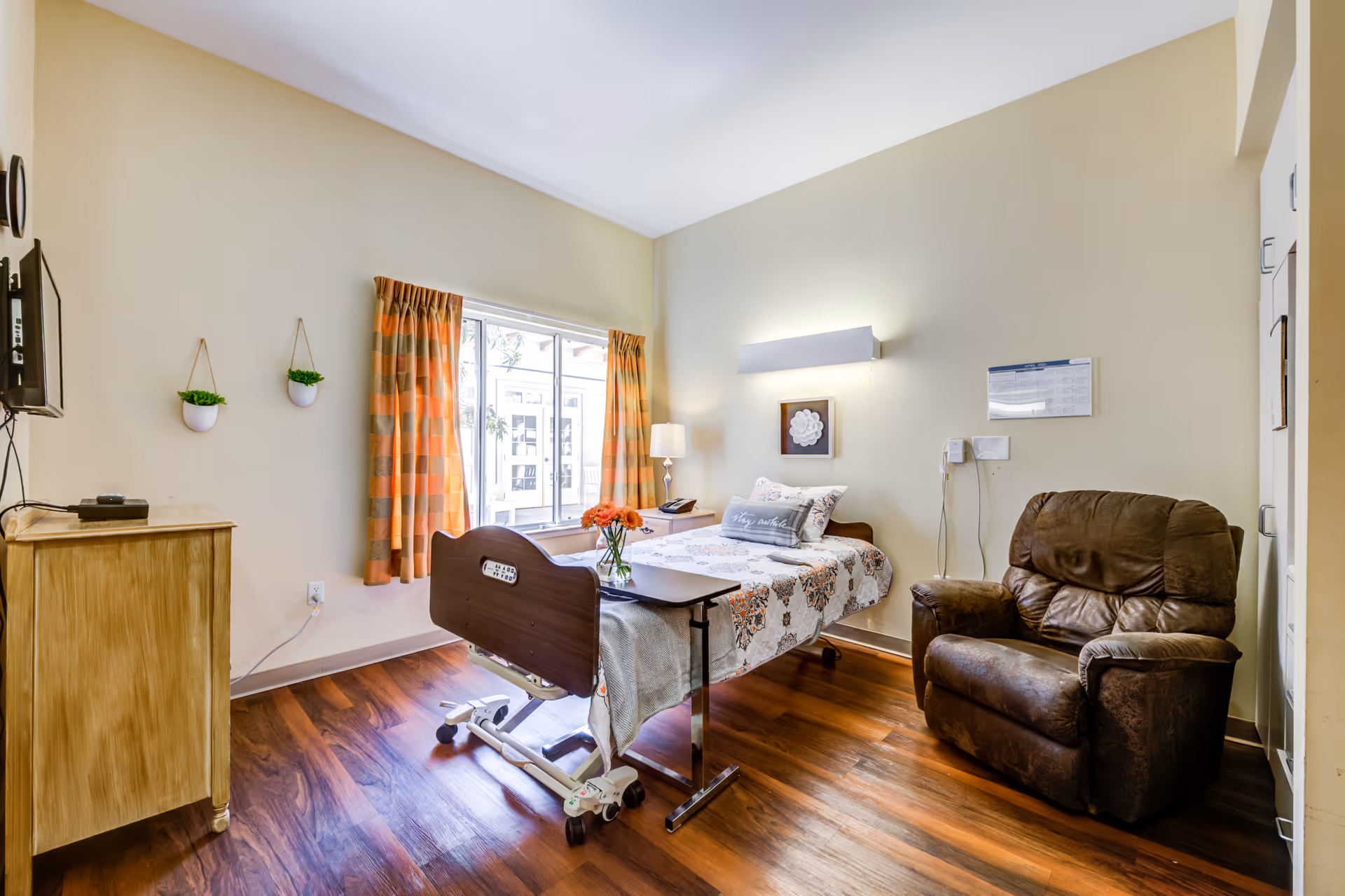 A senior living facility bedroom with a hospital-style adjustable bed covered in a floral quilt, a brown recliner chair, a wooden dresser with a TV mounted above it, two small hanging potted plants on the wall, a window with orange and beige checkered curtains, and a small table with a vase of flowers on the bed.