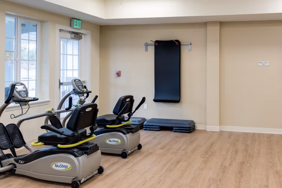 A small fitness room with two NuStep recumbent exercise bikes, a wall-mounted black exercise mat, and stacked step platforms on a wooden floor. There is a door with windows on the left side and an exit sign above it.