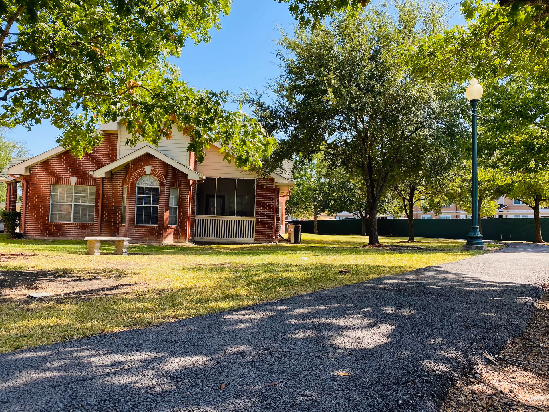 A single-story brick building with white trim surrounded by green grass and trees under a clear blue sky. There is a paved pathway leading past the building, a stone bench on the lawn, and a tall street lamp near the path.