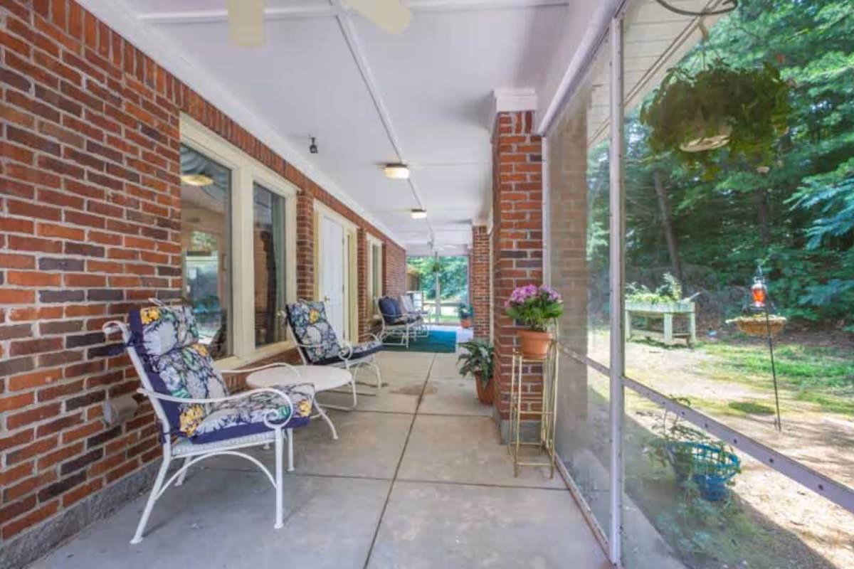 A covered outdoor patio area with brick walls and white-framed windows and doors. There are several cushioned chairs with floral patterns and a small white table. Potted plants and hanging greenery decorate the space. The patio overlooks a garden area with trees and a bird feeder visible through a screened enclosure.