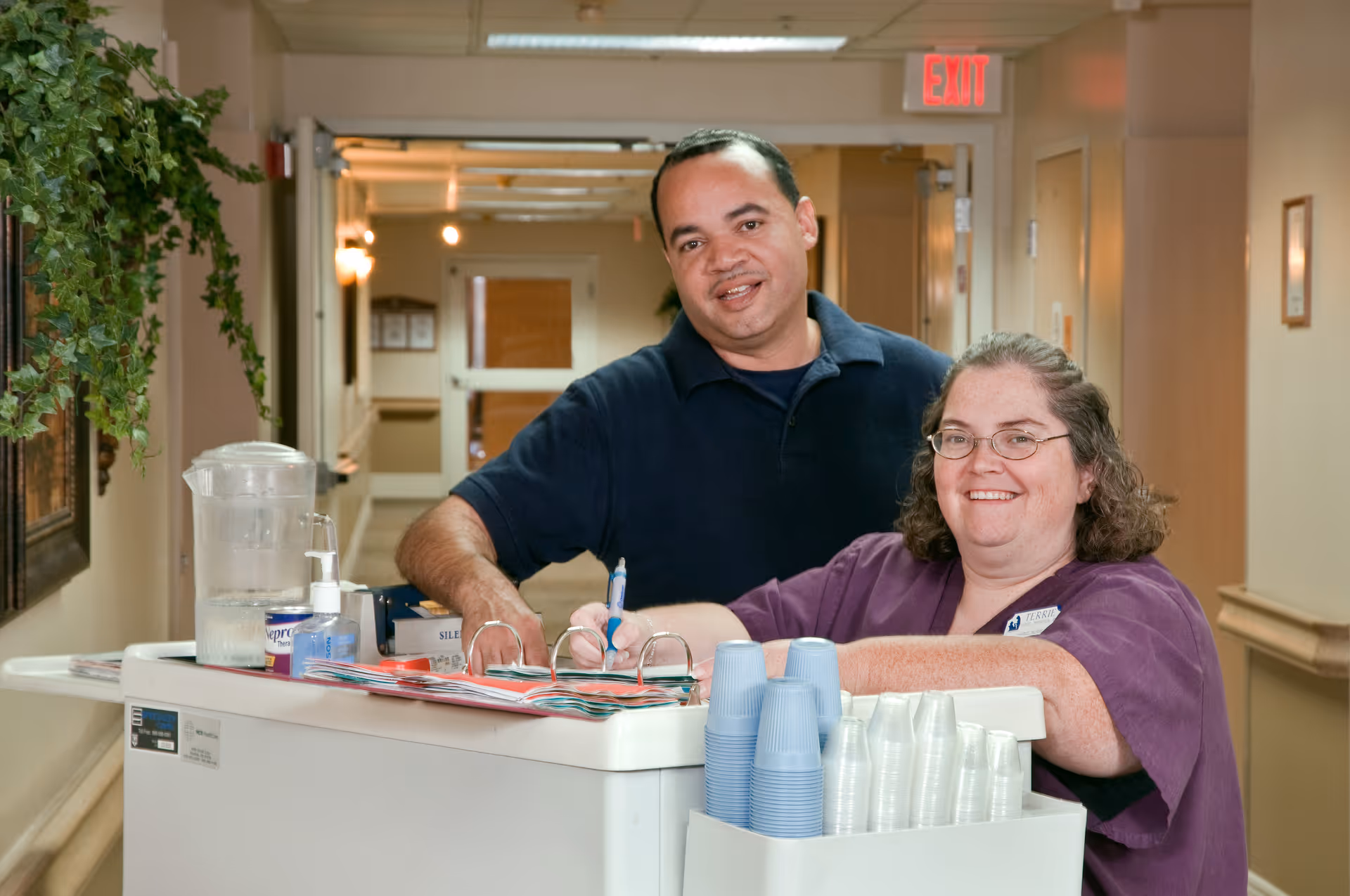 Two healthcare workers smiling in a hallway of a senior living facility. One is a man in a dark blue shirt and the other is a woman in purple scrubs with glasses, standing behind a cart with cups, a water pitcher, and paperwork.