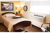 A tidy nursing facility bedroom with a made bed, bedside table and lamp, wall clock, window with air conditioner, chair, and framed artwork.