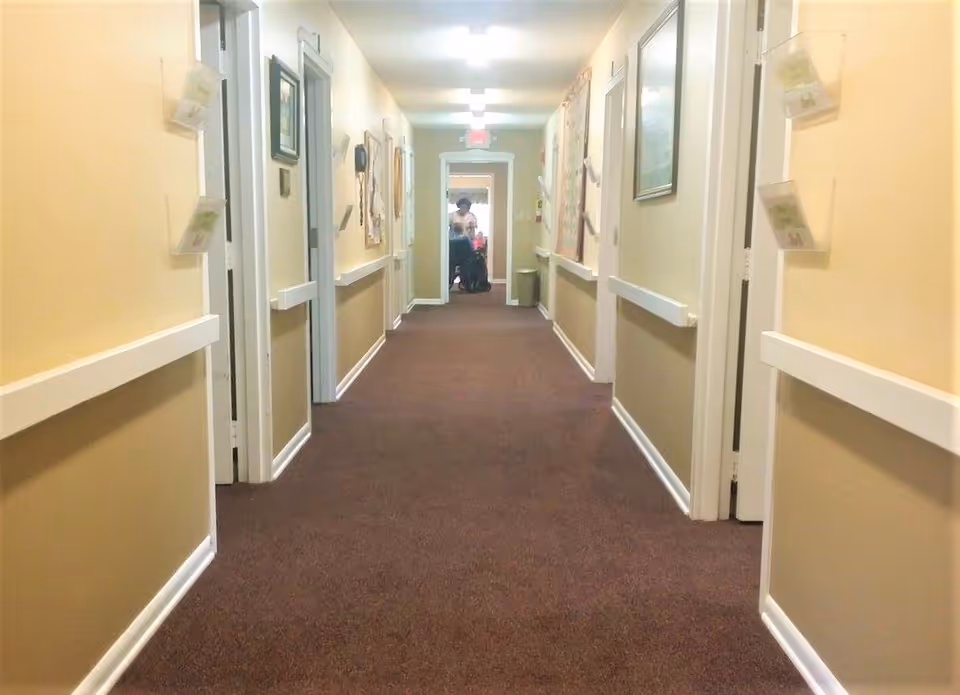 A long hallway in a senior living facility with beige walls and brown carpet. There are several doors on both sides, handrails along the walls, bulletin boards, and framed pictures. At the far end of the hallway, a person is visible, possibly in a wheelchair.