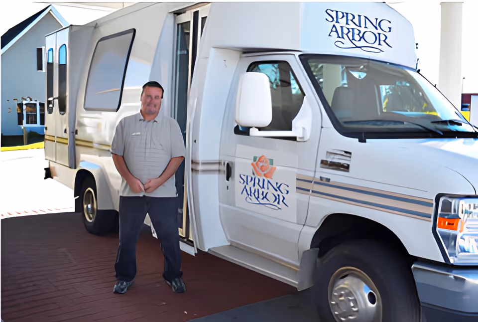 A Spring Arbor shuttle van parked at a facility entrance with a staff member standing in the open doorway.