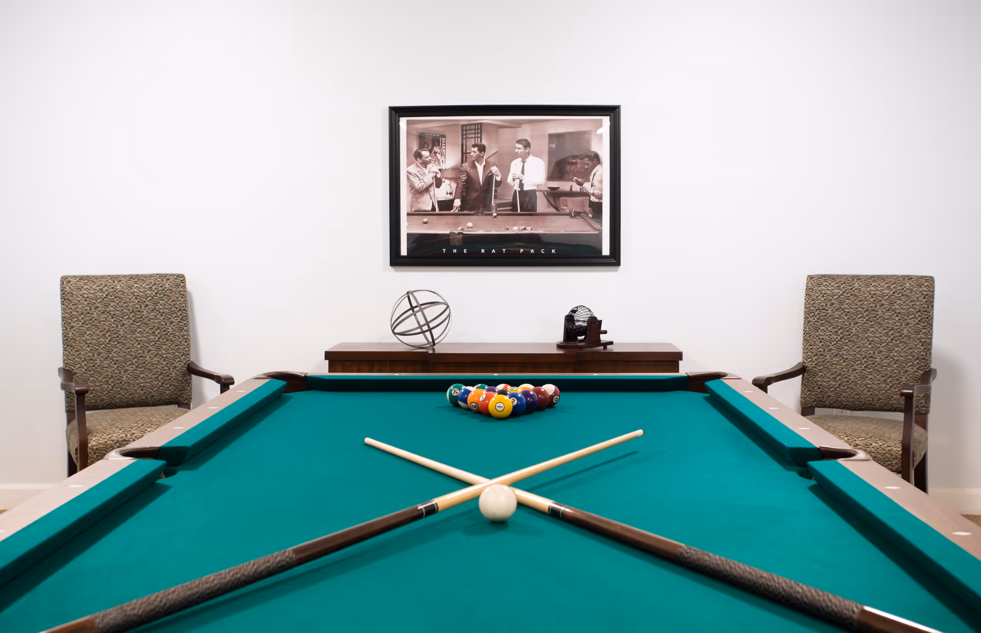 A pool table with two crossed pool cues and a rack of billiard balls set up for a game. Behind the pool table are two upholstered armchairs and a wooden console table with decorative items. A framed black and white photo of four men playing pool hangs on the white wall above the console table.