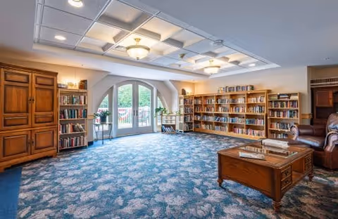 A spacious senior living common area with blue patterned carpet, wooden bookshelves filled with books, a wooden coffee table with books on it, a leather armchair, and large arched glass doors leading outside. The ceiling has recessed lighting and decorative panels.