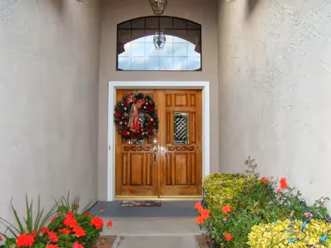 Wooden double front doors decorated with a Christmas wreath beneath an arched transom window, set in a sheltered entryway with flowering shrubs.