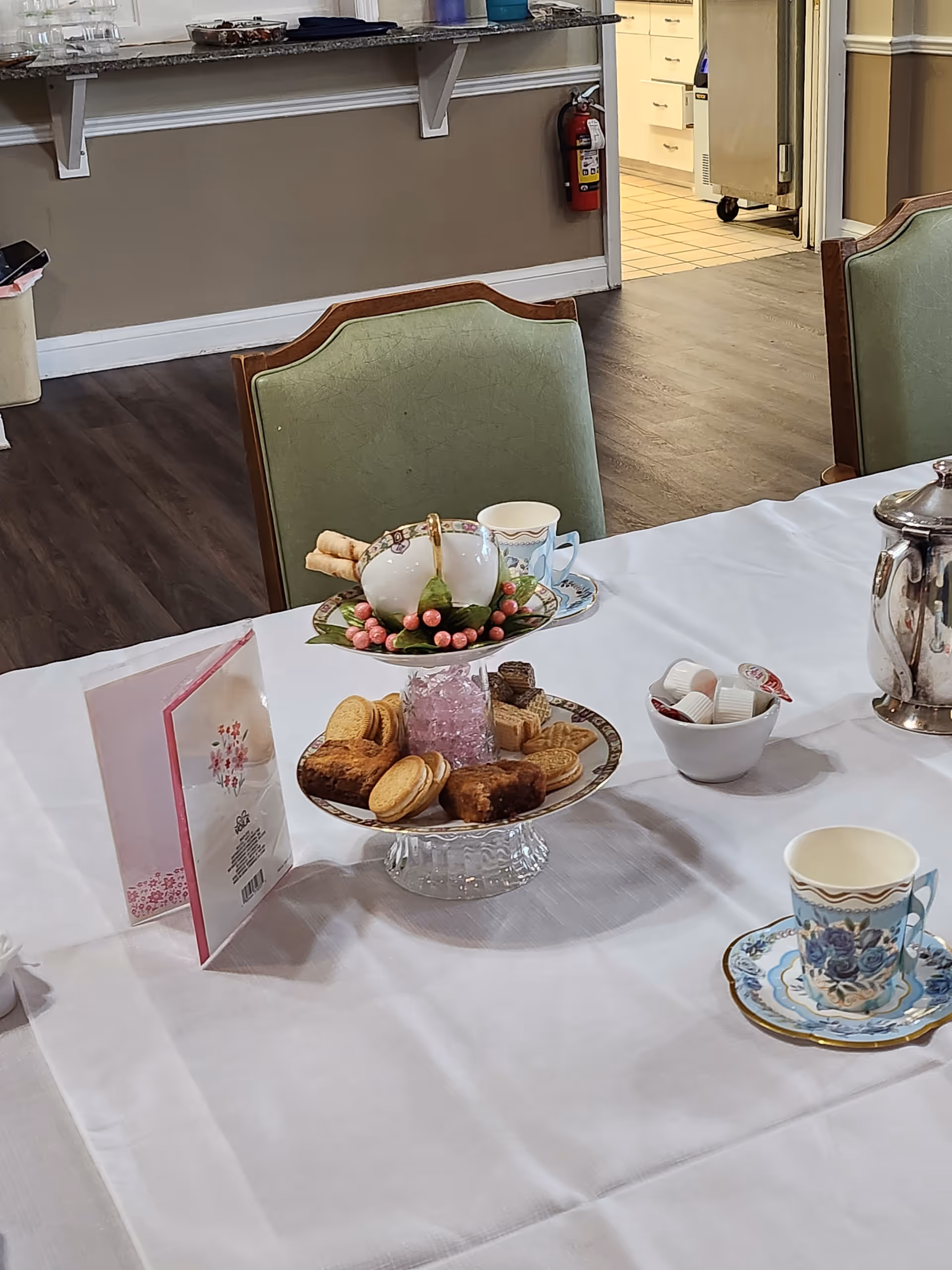A table set for tea with a white tablecloth, featuring a two-tiered serving tray with assorted cookies and sweets, a floral teapot, teacups with saucers, a small bowl with creamers, and a silver teapot. In the background, there are green upholstered chairs, a counter with items on it, and a view into a kitchen area with tiled flooring.