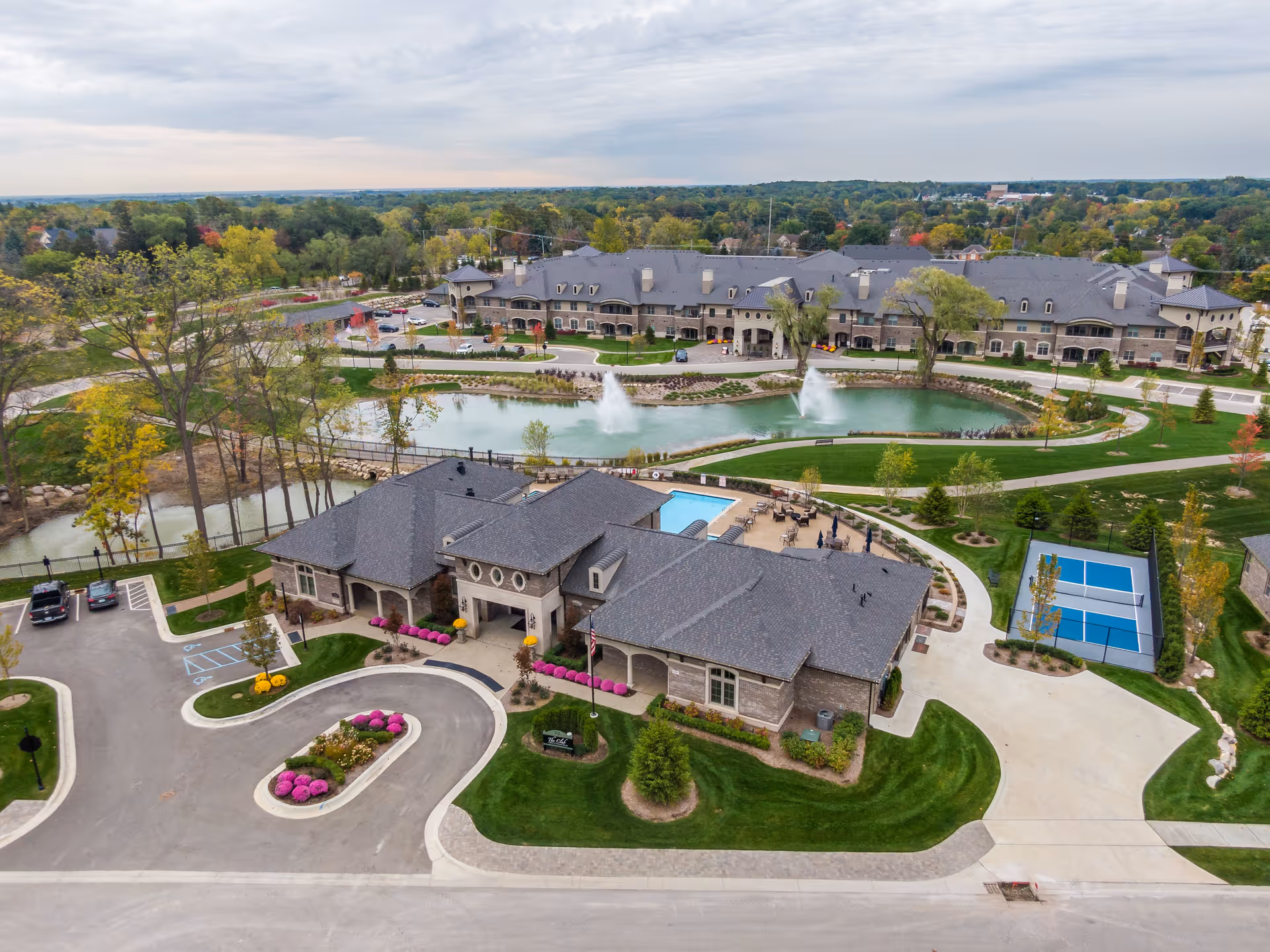 Aerial view of Villages at Flourish Collection senior living facility showing multiple buildings with gray roofs, a small pond with two water fountains, a swimming pool, a tennis or pickleball court, landscaped green lawns, and parking areas surrounded by trees and a cloudy sky.