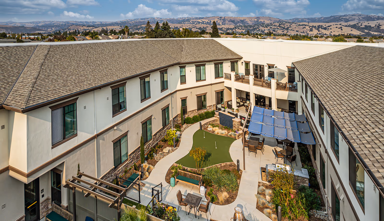 Aerial view of a two-story senior living courtyard featuring seating areas, a small putting green, shaded patio, and surrounding balconies with hills in the distance.