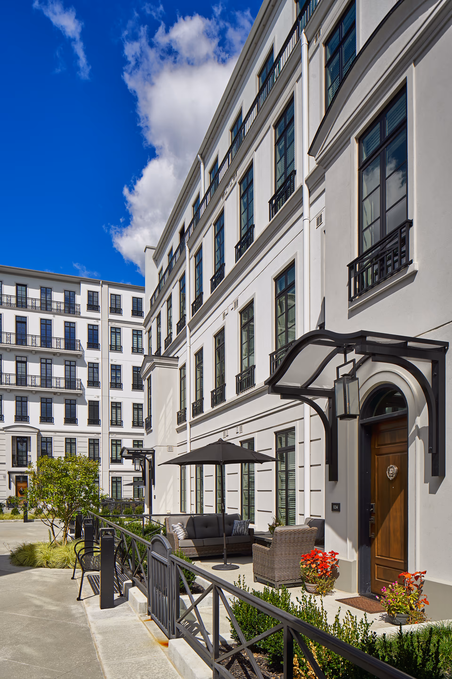 Outdoor seating area in front of a multi-story white building with large windows and black railings. The area includes a patio with a sofa, chairs, an umbrella, potted plants, and a wooden door with a decorative awning. The sky is blue with some clouds.