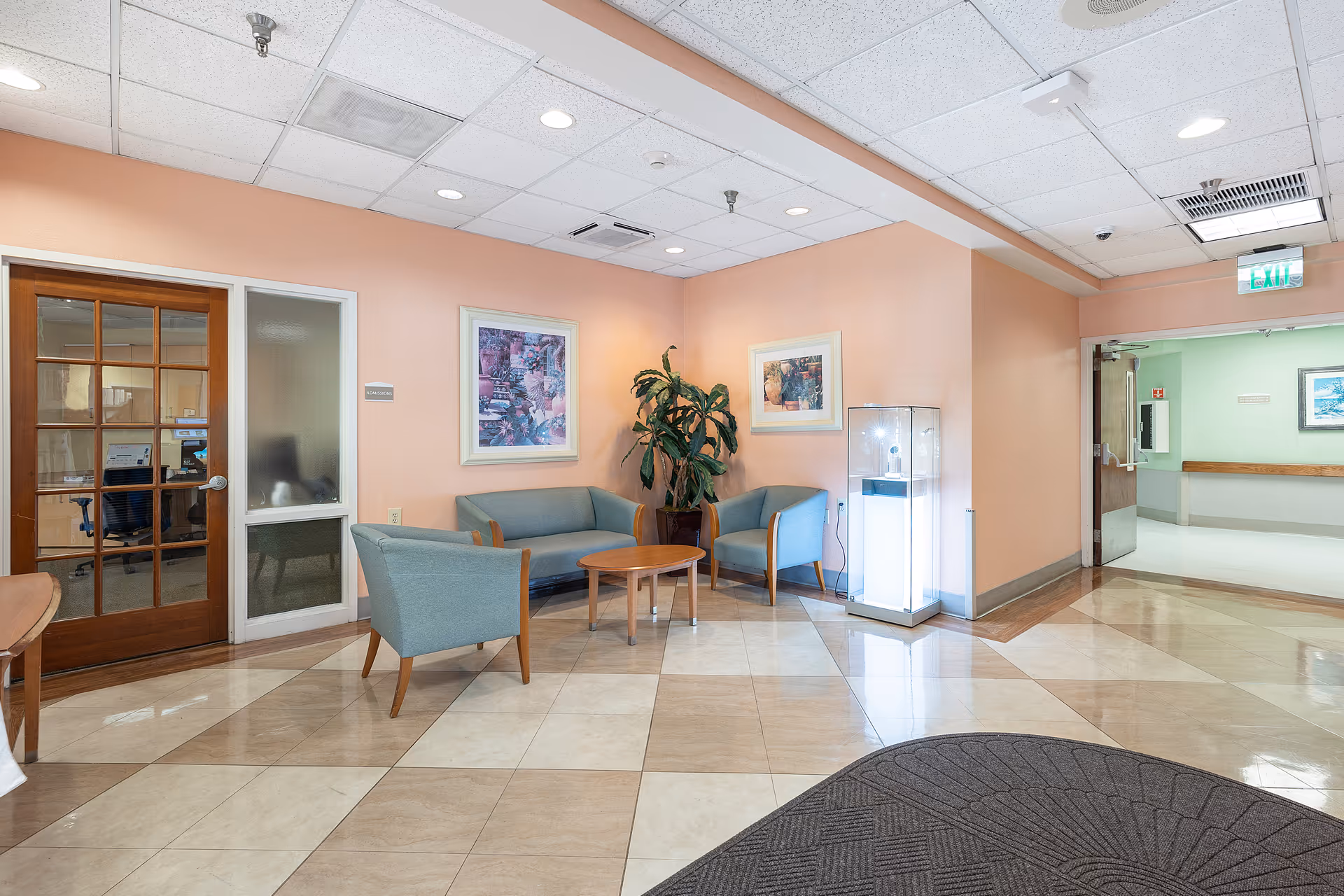 Lobby seating area with blue upholstered chairs and sofa around a wooden coffee table, artwork on peach walls, and tiled floor.