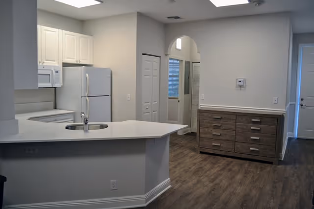 Interior view of a kitchen area with white cabinets, a white refrigerator, a microwave, and a countertop with a built-in sink. The kitchen opens into a hallway with wooden flooring and a wooden dresser against the wall. There is a white door at the end of the hallway.