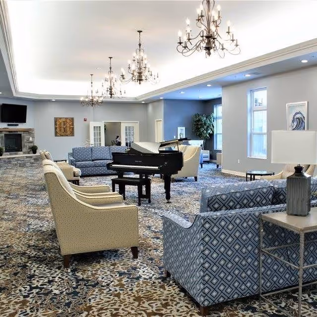 A spacious and well-lit living room area in an assisted living facility featuring multiple upholstered armchairs and sofas arranged around a black grand piano. The room has patterned carpet flooring, several elegant chandeliers hanging from the ceiling, large windows allowing natural light, a stone fireplace with a mounted TV above it, and decorative wall art.