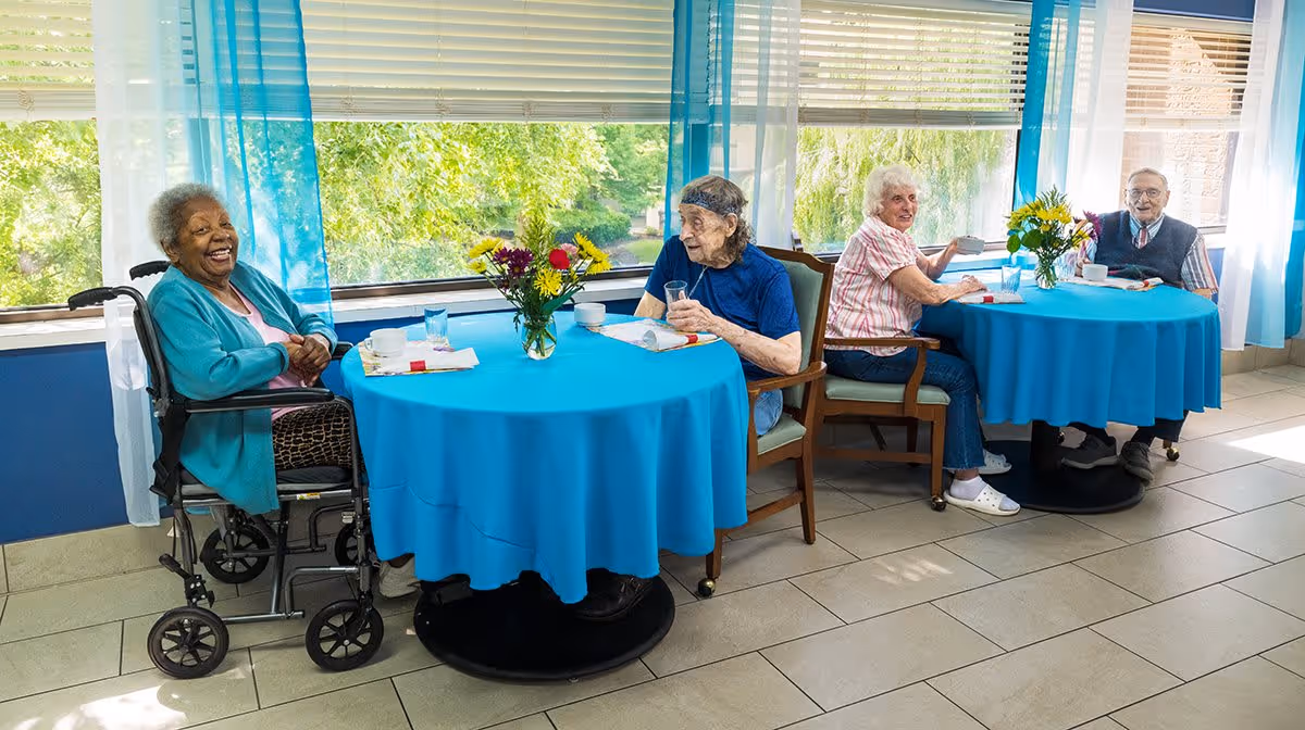 Four elderly people sitting at two round tables covered with blue tablecloths in a bright room with large windows and blue curtains. They are enjoying drinks and snacks, with flower vases on each table. One person is in a wheelchair, and the others are seated on chairs.