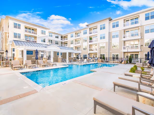 Outdoor swimming pool area surrounded by lounge chairs and umbrellas, with a multi-story residential building in the background under a partly cloudy blue sky.