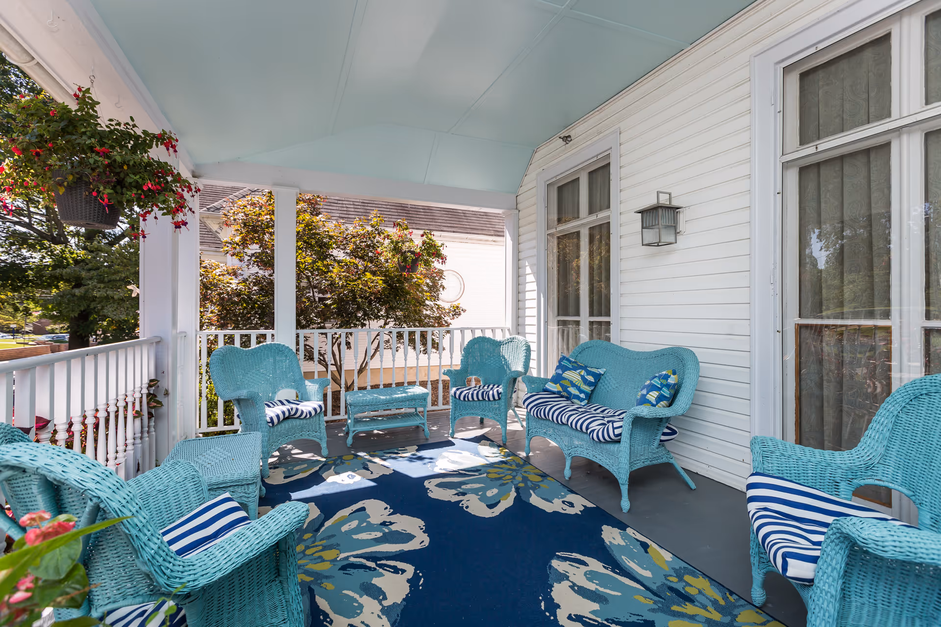 Covered outdoor porch area with light blue wicker chairs and loveseat, striped and patterned cushions, a floral area rug, white railing, hanging flower basket, and white siding walls with windows and a wall-mounted lantern.