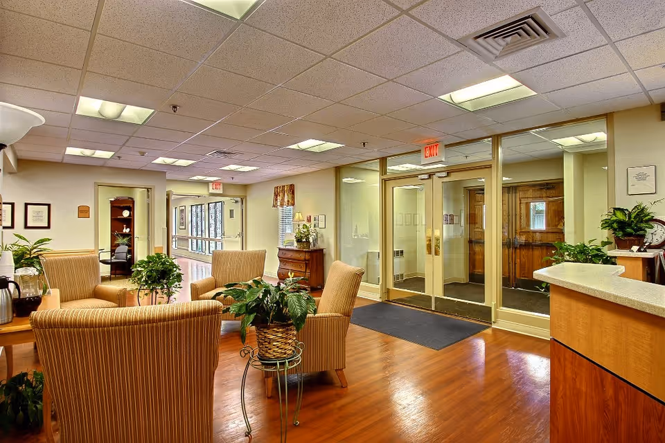 A well-lit seating area in a healthcare facility with four striped armchairs arranged around a small table with a potted plant. The room has wooden flooring, several green plants, and a reception desk on the right. Glass double doors with an exit sign above lead to another area, and framed pictures and certificates hang on the walls.